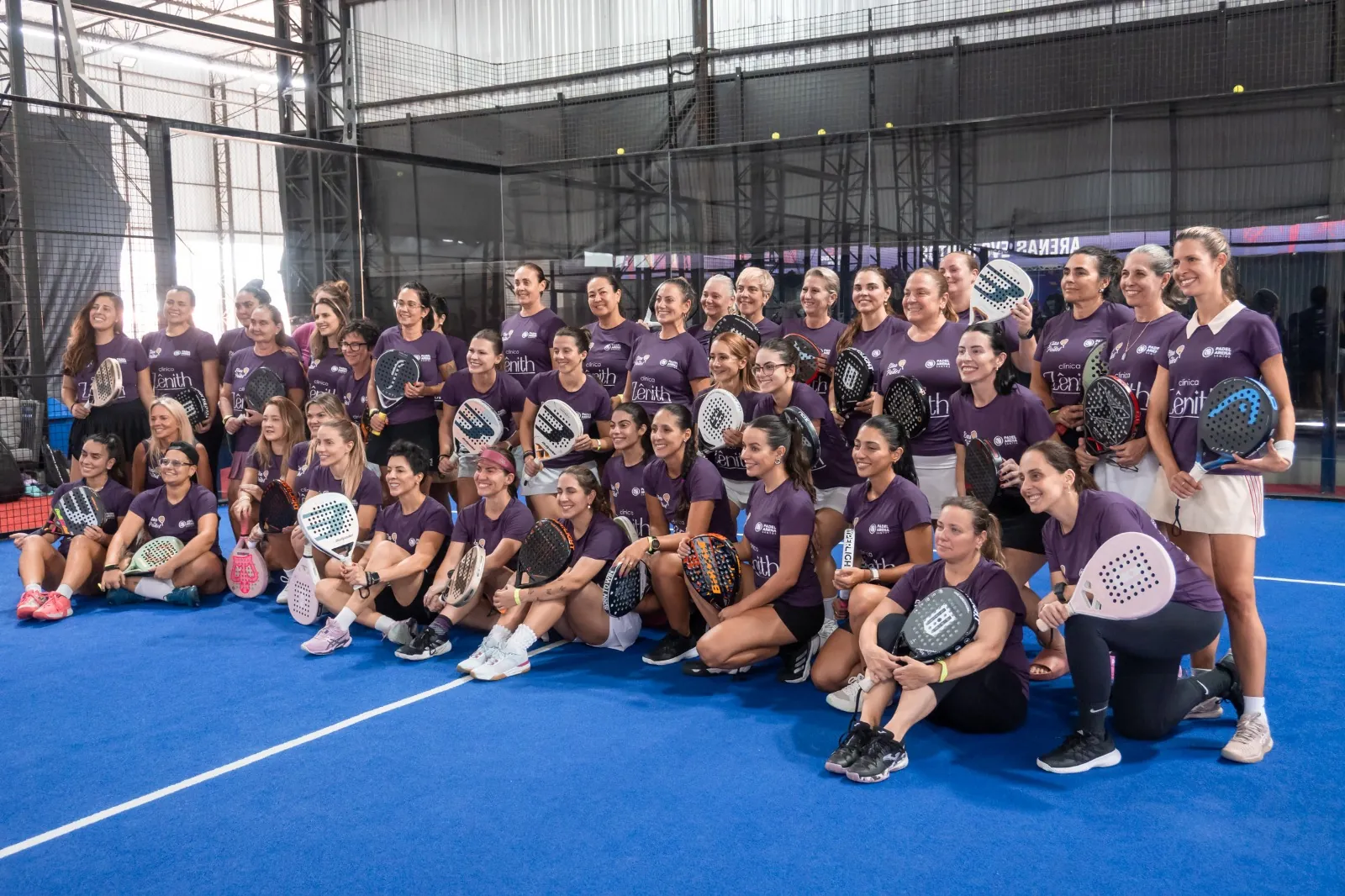 Turma feminina de camiseta roxa reunida com as raquetes na quadra azul do Padel Center